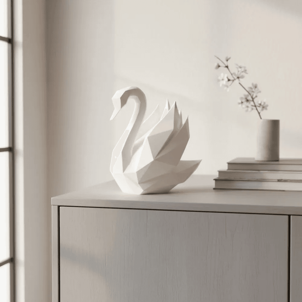 Hvid low poly svanefigur på en lys skænk ved et vindue, med bøger og en vase med gren