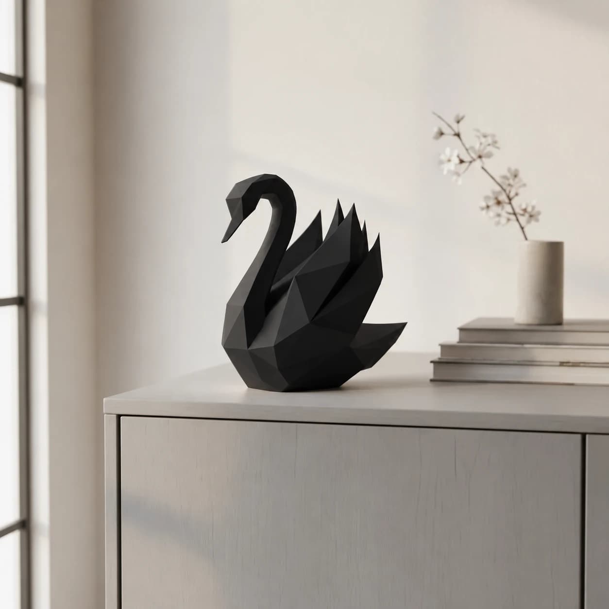Sort low poly svanefigur i mat sort kul på en lys skænk ved et vindue, med bøger og en vase med gren