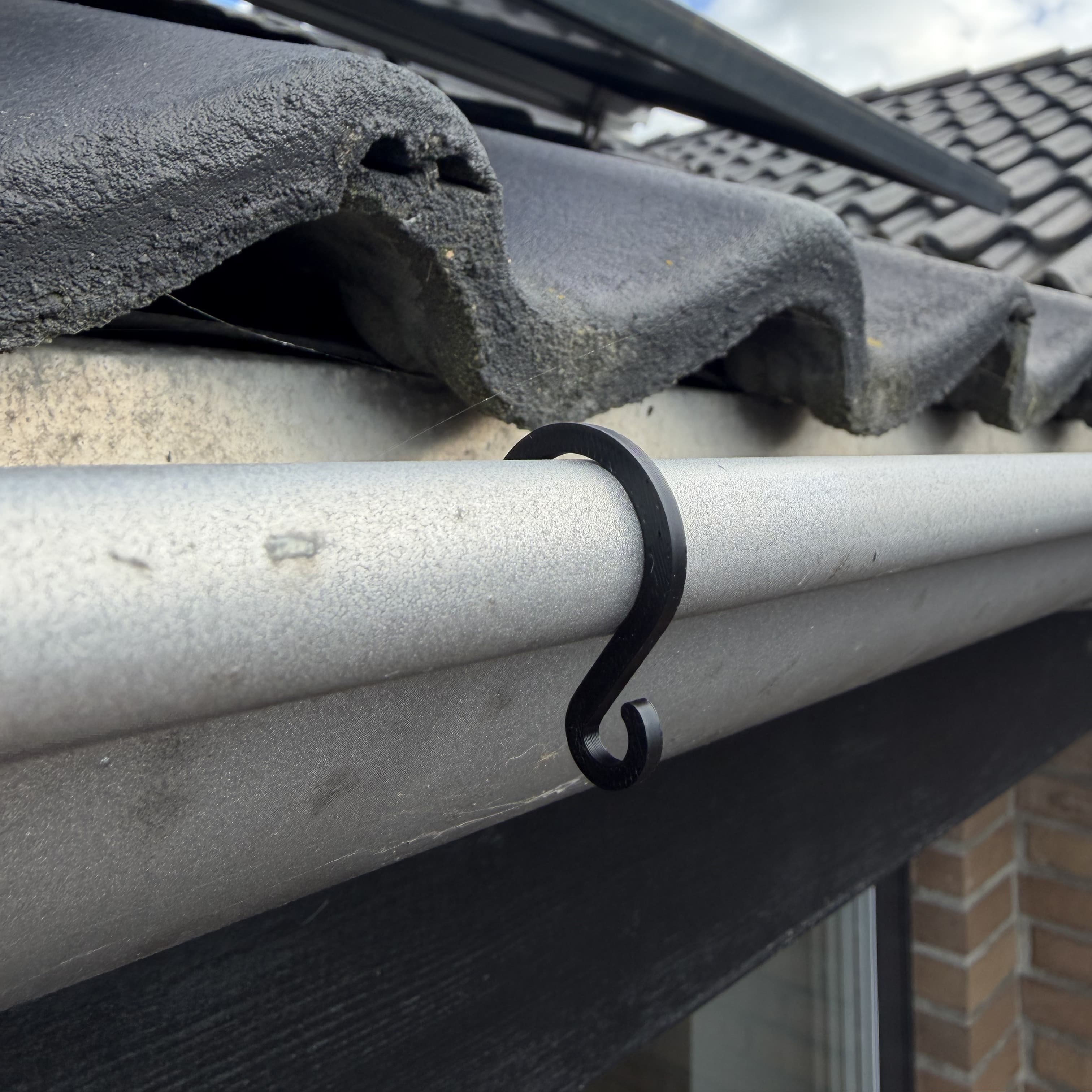 Sort 3D-printet S-formet clips på vulsten af en halvrund grå metaltagrende under betontagsten